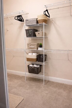 A white metal shelving unit with baskets and bags, labeled 'Clothes' on the middle shelf.