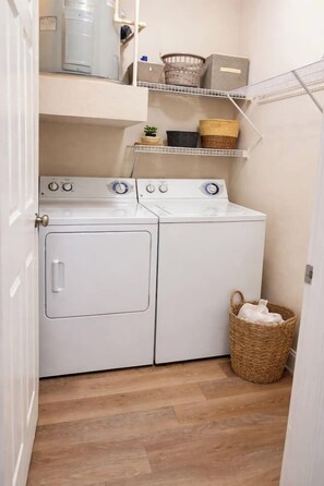 A laundry room with a front-loading washer and dryer, shelves above with baskets, and a wicker basket on the floor. - Heart & Soul Texas wParking WIFI Galleria MedCenter NRG (Houston)