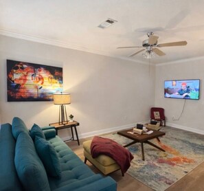 A living room with a sofa, a coffee table, a television, a ceiling fan, and a painting on the wall.