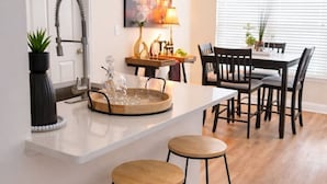 Una cocina con encimera blanca, dos taburetes, una mesa de comedor con sillas y un cuadro en la pared.
