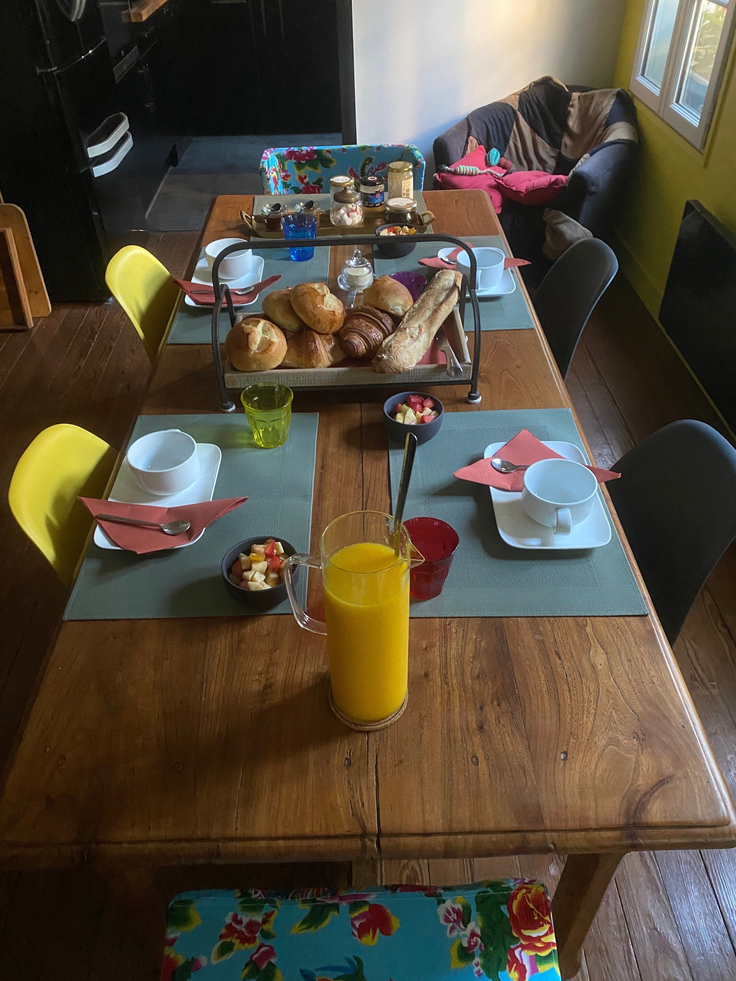 A dining table set with breakfast items, including pastries, a pitcher of orange juice, and a bowl of fruit.