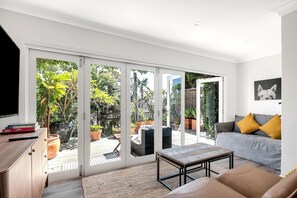 Ein modernes Wohnzimmer mit einer Couch, einem Couchtisch und einem Fernseher. Ein Schiebeglasfenster führt auf eine Terrasse mit Pflanzen und Gartenmöbeln.