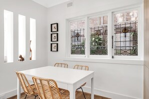 Ein Essbereich mit weißem Tisch und Korbstühlen, einem Fenster mit Buntglas und gerahmten Bildern an der Wand.