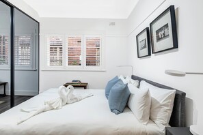 Ein modernes Schlafzimmer mit Bett, Kissen, einem Nachttisch, einem Fenster mit Jalousien und gerahmten Bildern an der Wand.