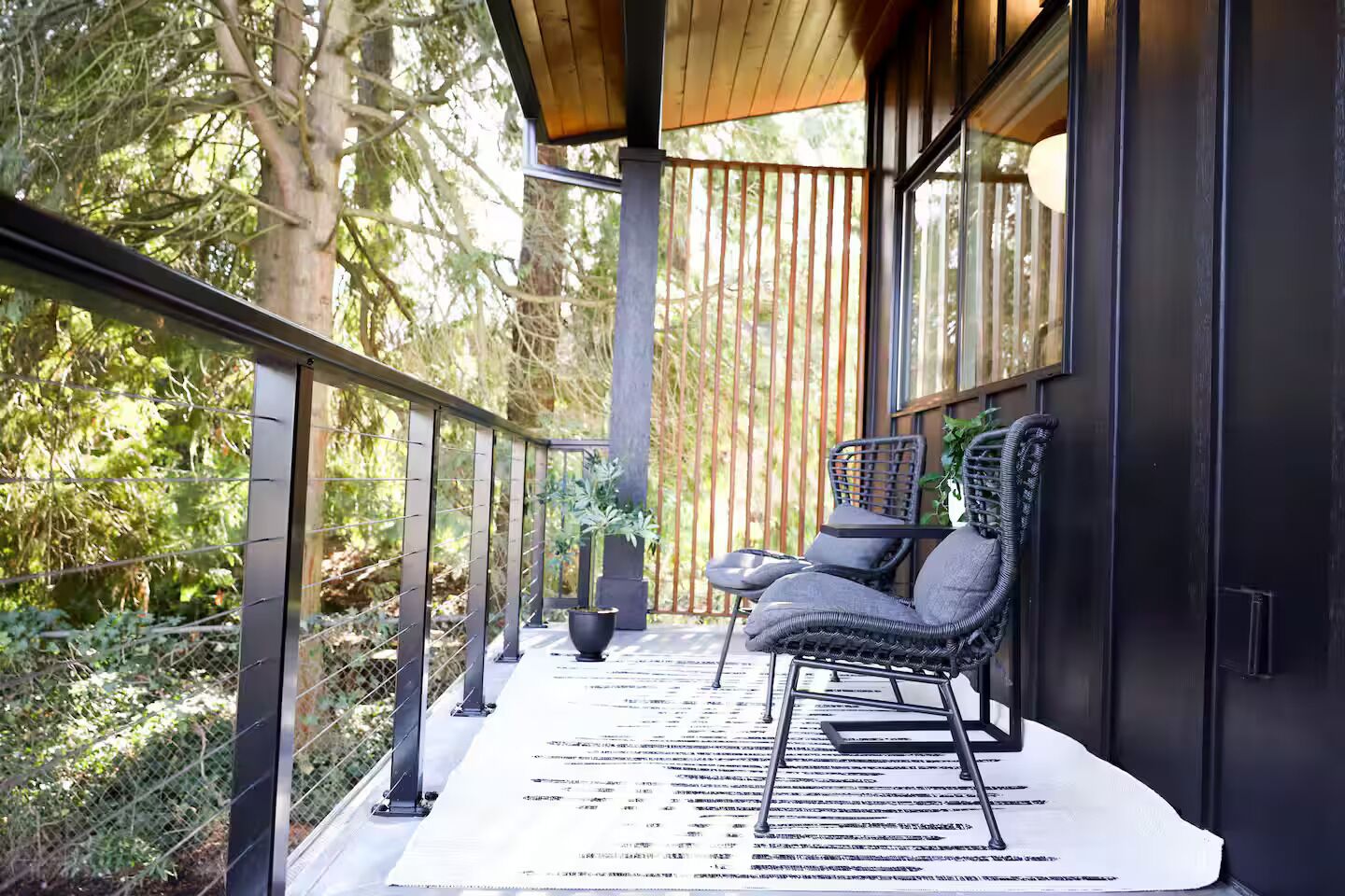 A balcony with a chair and a table, surrounded by a wooden railing and glass walls, overlooking a forested area.