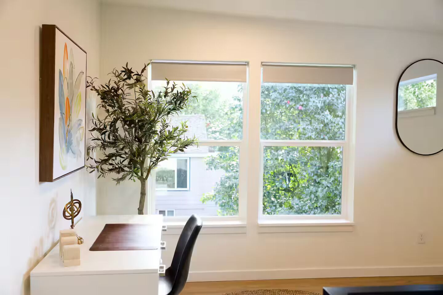 Une pièce avec un bureau, deux fenêtres, une plante, un miroir et un tableau accroché au mur.