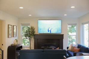 A living room with a flat-screen TV mounted above a fireplace, a dark-colored sofa, and a potted plant. - Luxury 3BDR Home - Near PDX - Balcony & Fireplace (Vancouver)