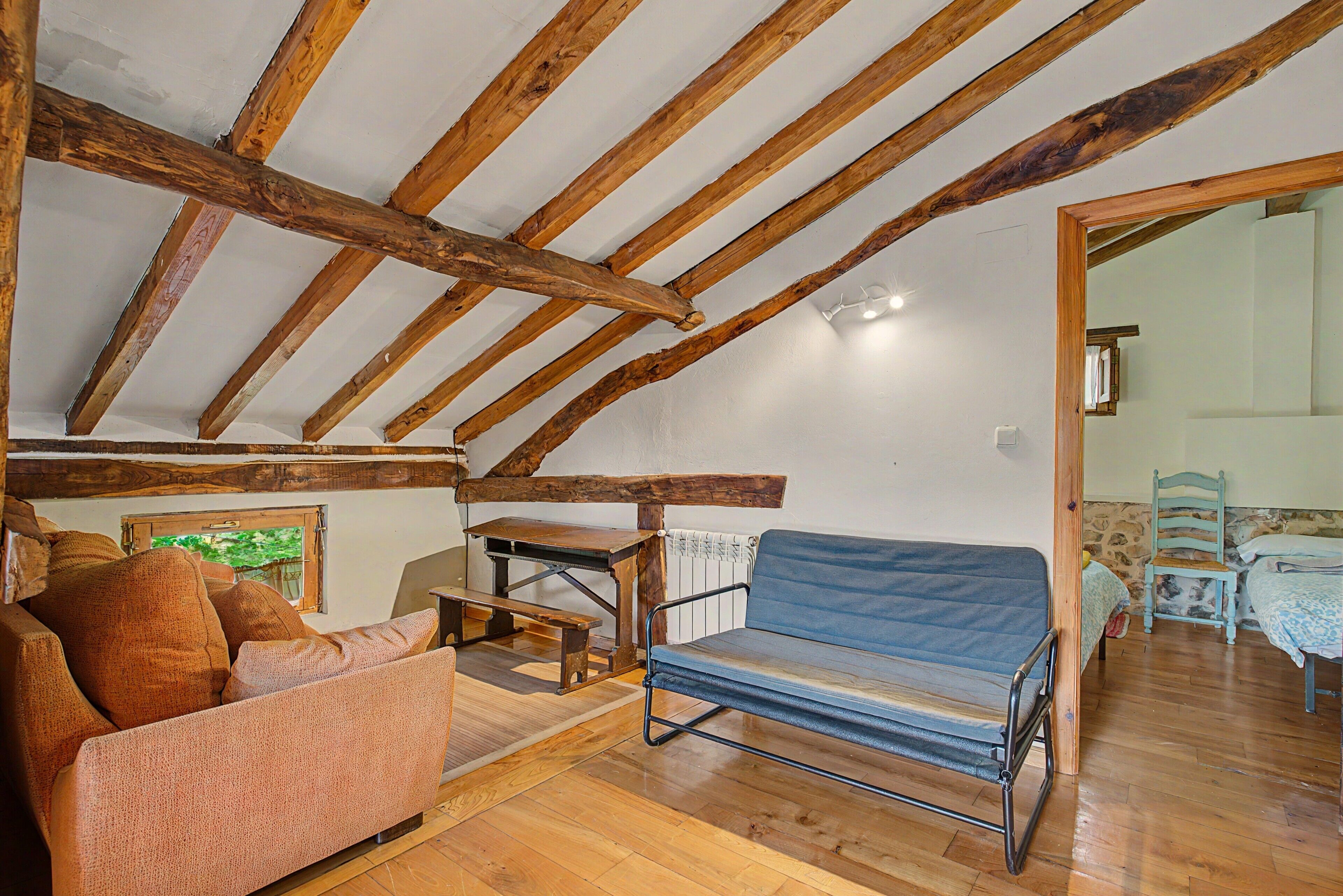 A rustic attic room with exposed wooden beams, a blue cushioned bench, a wooden table, and a sofa.