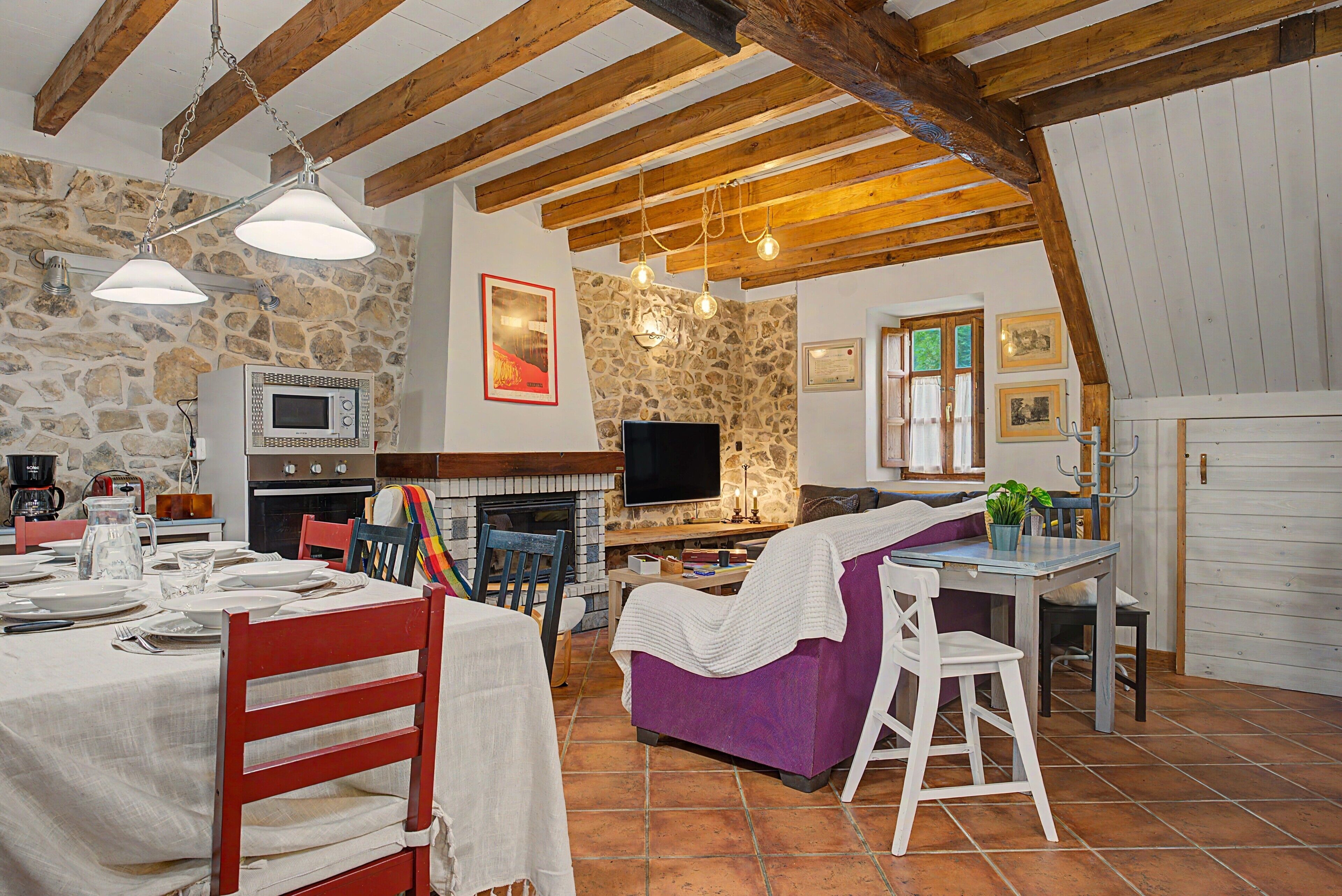 Un interior rústico con vigas de madera a la vista, pared de piedra, chimenea, televisor y comedor listo para una comida.