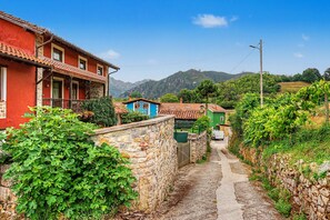 Un étroit sentier de pierre mène à des maisons traditionnelles aux toits de tuiles, entourées d’une végétation luxuriante et avec des montagnes en toile de fond.