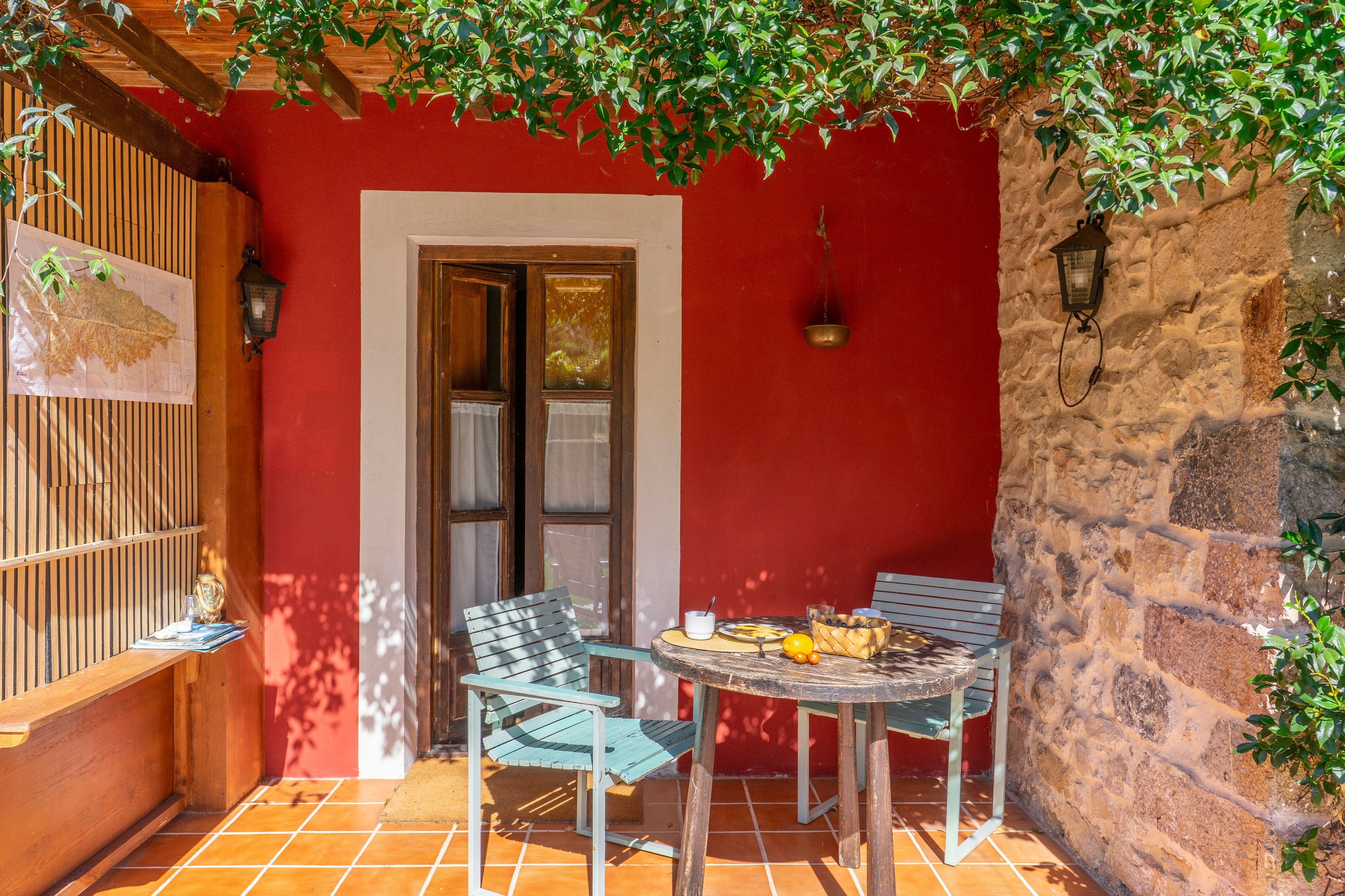 Un patio con un muro rojo, una puerta de madera, dos sillas, una mesa con frutas y un mapa en la pared.