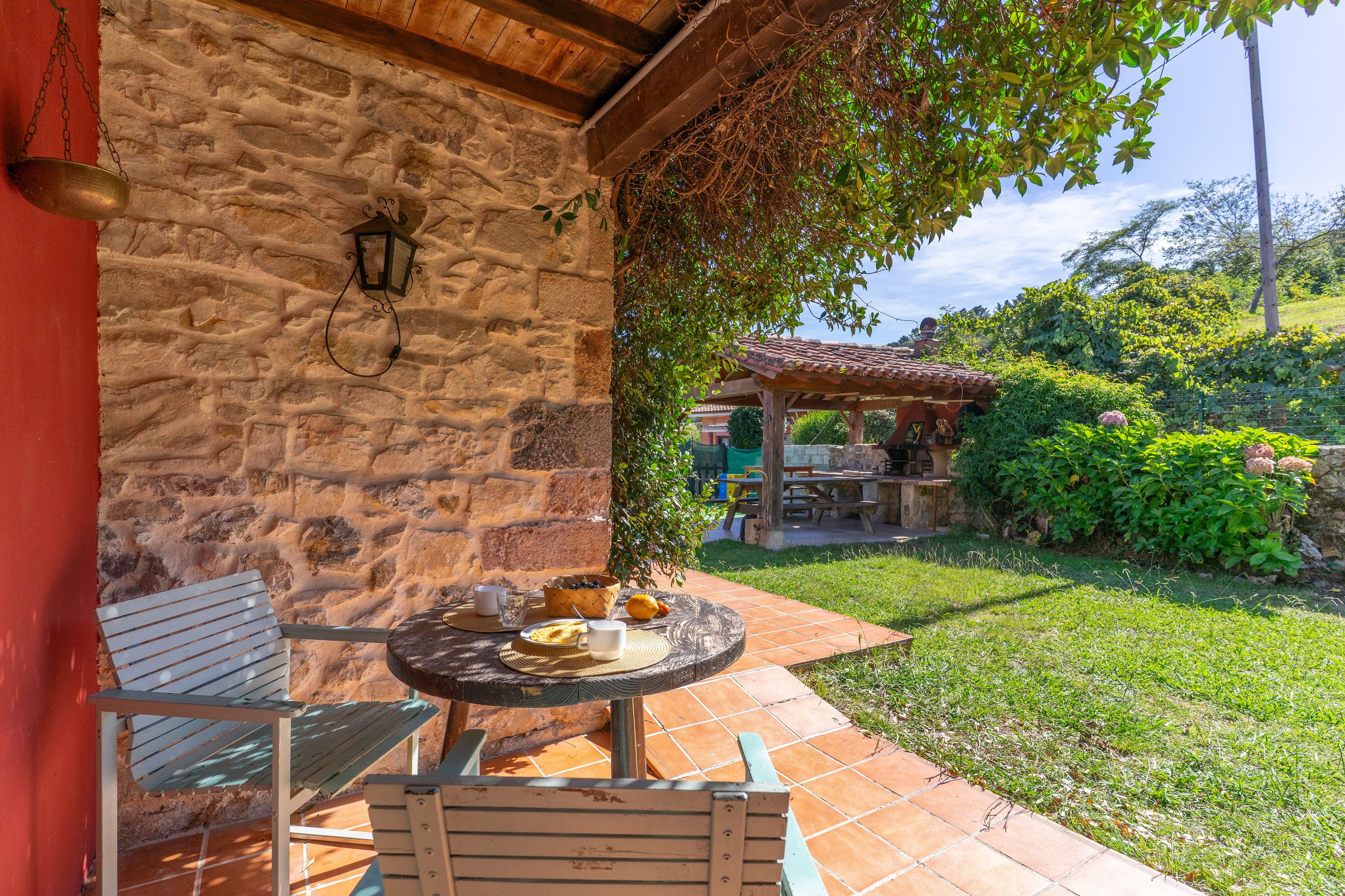 Un muro de piedra con un farol colgante, un banco de madera y una mesa con una cesta de fruta y una taza.