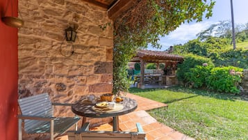 Un muro de piedra con un farol colgante, un banco de madera y una mesa con una cesta de fruta y una taza.