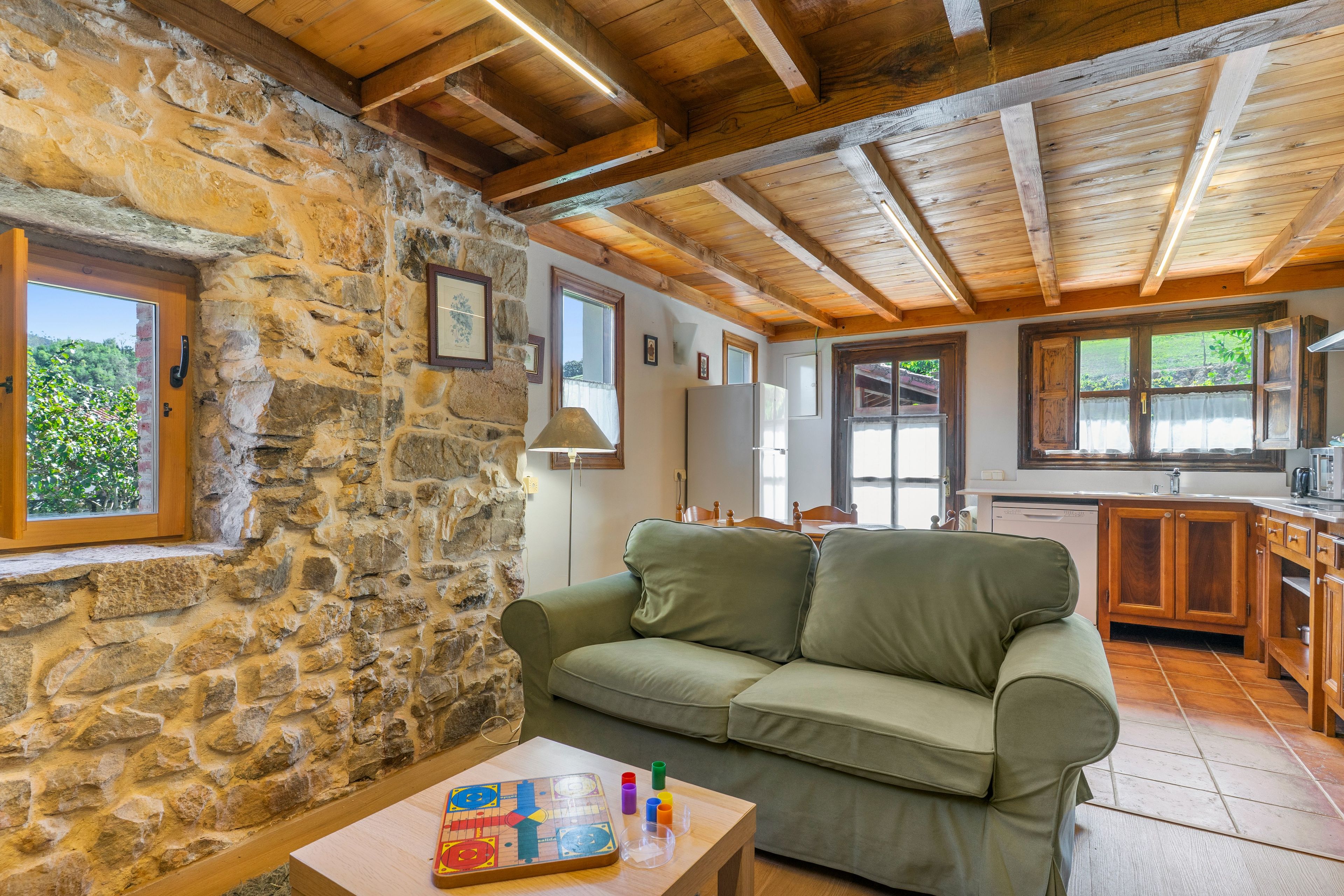 Una habitación acogedora con pared de piedra, vigas de madera en el techo, un sofá verde, una mesa con un juego de mesa y una zona de cocina con armarios de madera y un ventanal.