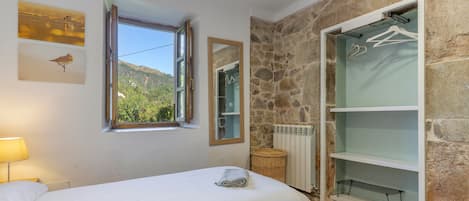Un dormitorio con cama, ventana con vista a un bosque, pared de piedra y armario con perchas.