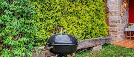 Una parrilla negra con tapa, ubicada en un área cubierta de césped con un muro de piedra y un seto al fondo.