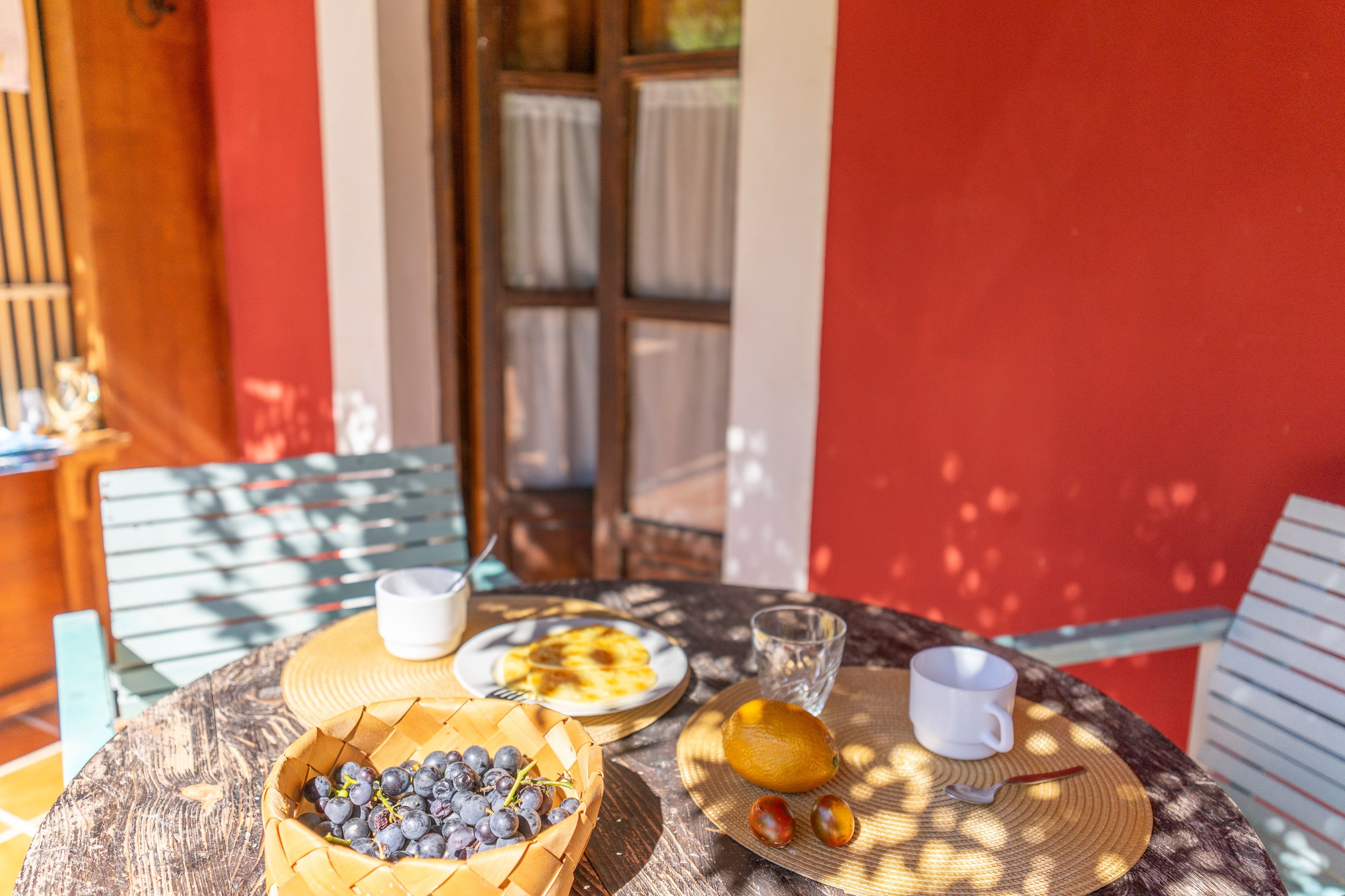 Une table dressée avec un panier de raisins, une assiette de nourriture, un verre d’eau et une tasse, le tout installé à l’extérieur, avec un mur rouge et une fenêtre en arrière-plan.