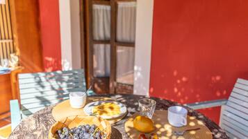 Une table dressée avec un panier de raisins, une assiette de nourriture, un verre d’eau et une tasse, le tout installé à l’extérieur, avec un mur rouge et une fenêtre en arrière-plan.