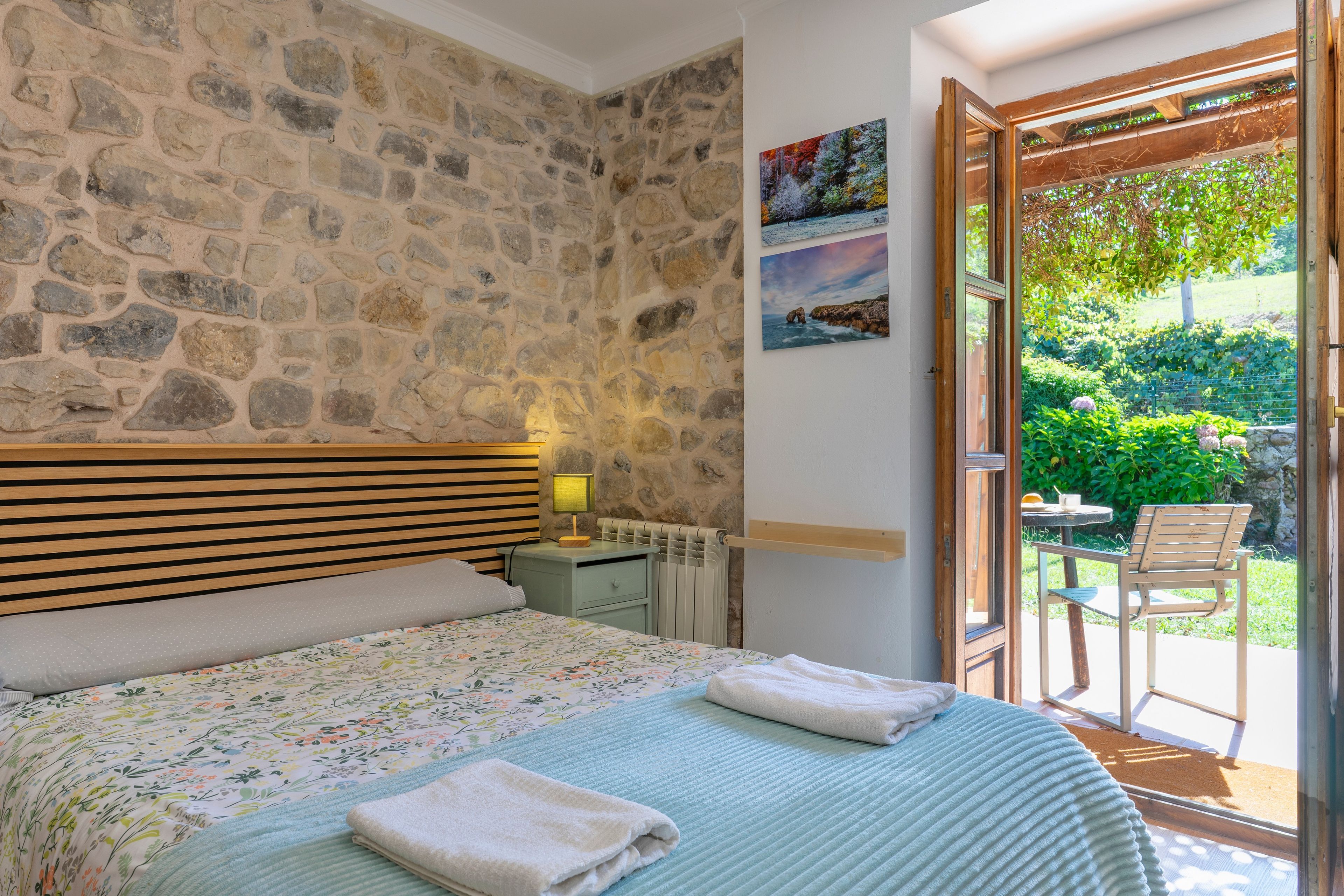 Un dormitorio con pared de piedra, cabecero de madera, cama con colcha floral, mesita de noche con lámpara y vista a un jardín a través de una puerta abierta.