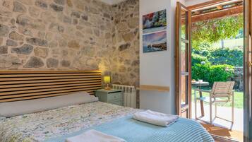 Un dormitorio con pared de piedra, cabecero de madera, cama con colcha floral, mesita de noche con lámpara y vista a un jardín a través de una puerta abierta.