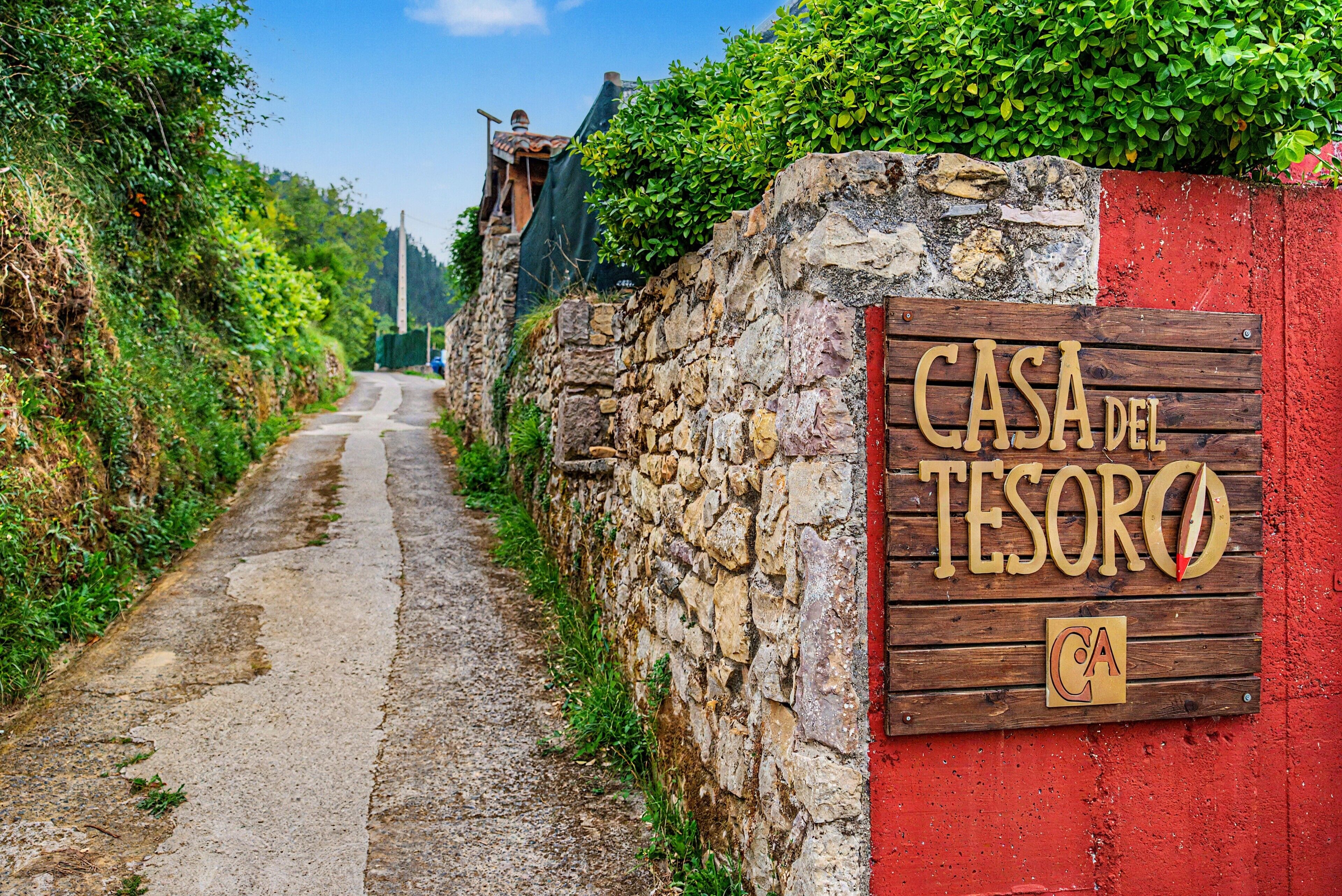 Una pared de piedra rústica con un letrero de madera que dice 'CASA DEL TESORO' y un estrecho sendero de tierra que se adentra en la distancia.