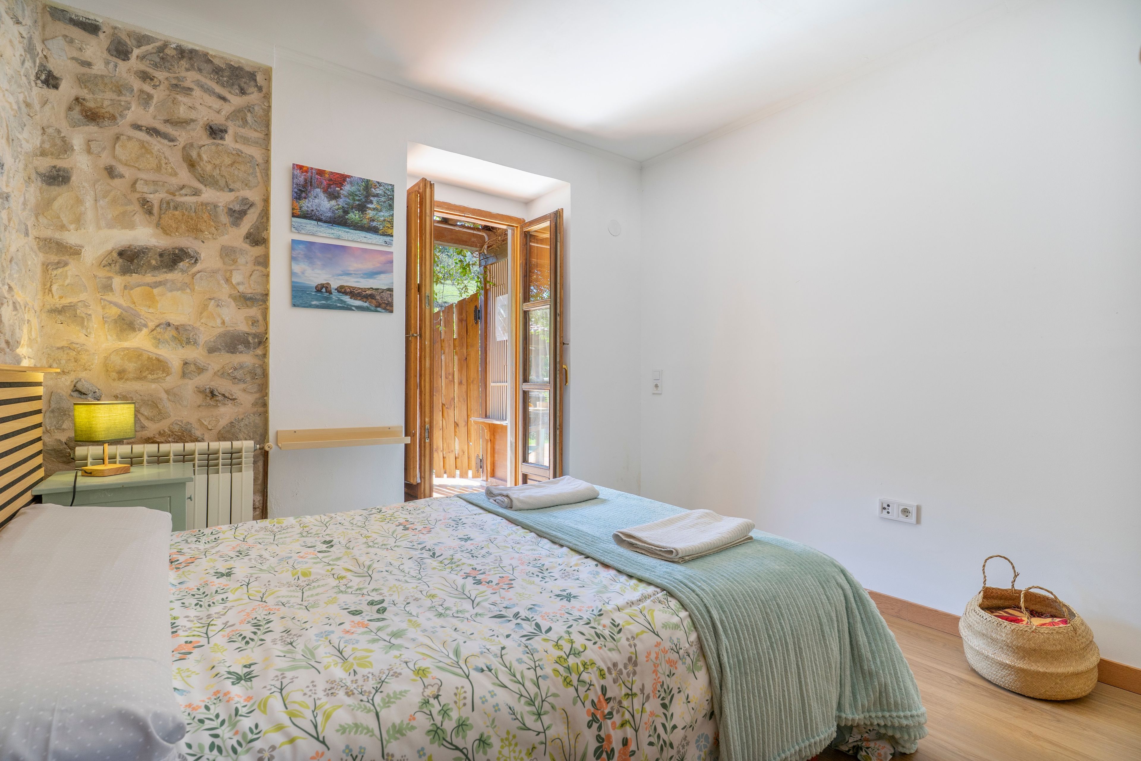 A bedroom with a bed, a wooden headboard, a stone wall, a painting, and a view through an open door.
