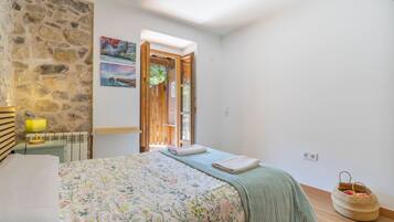 A bedroom with a bed, a wooden headboard, a stone wall, a painting, and a view through an open door.