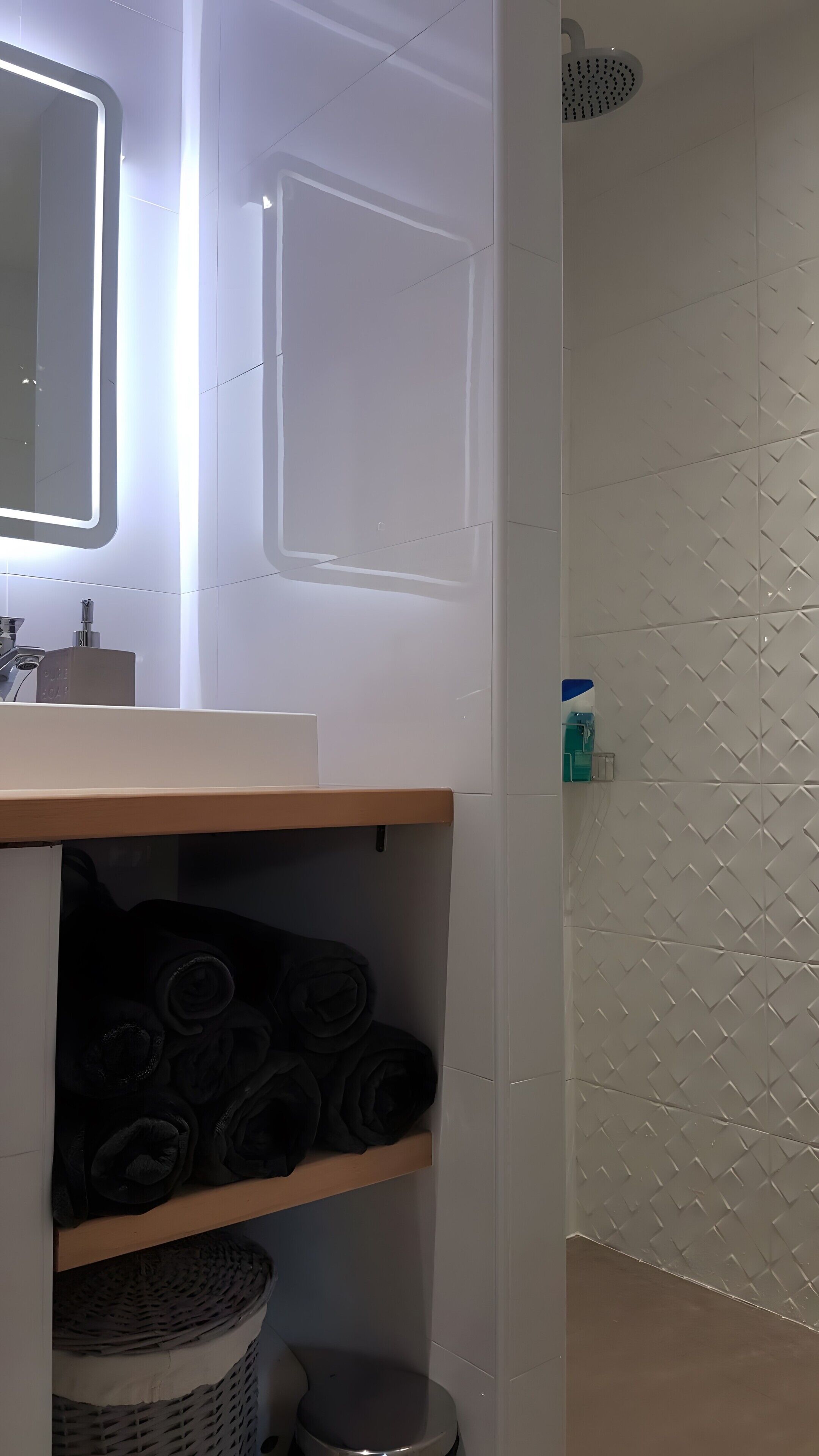 A bathroom with a white sink, mirror, and shower. There are shelves with rolled towels and a trash bin.