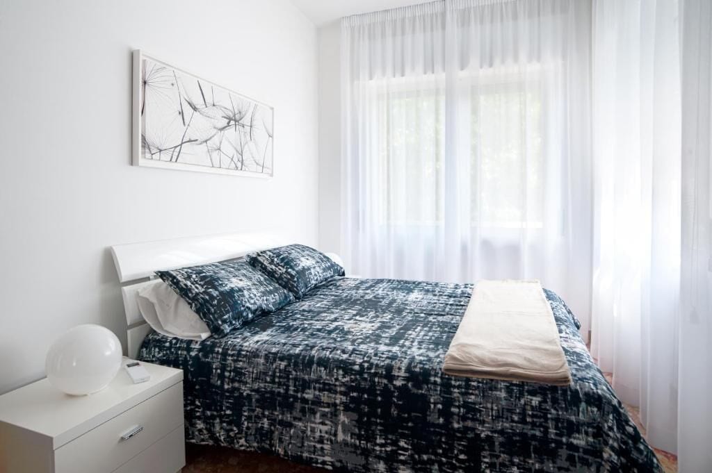 Une chambre à coucher avec un lit, une table de chevet, une lampe et une fenêtre avec des rideaux légers.
