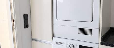 A compact, white stacked washer and dryer unit with a control panel, located in a room with a tiled floor and a door to the left.