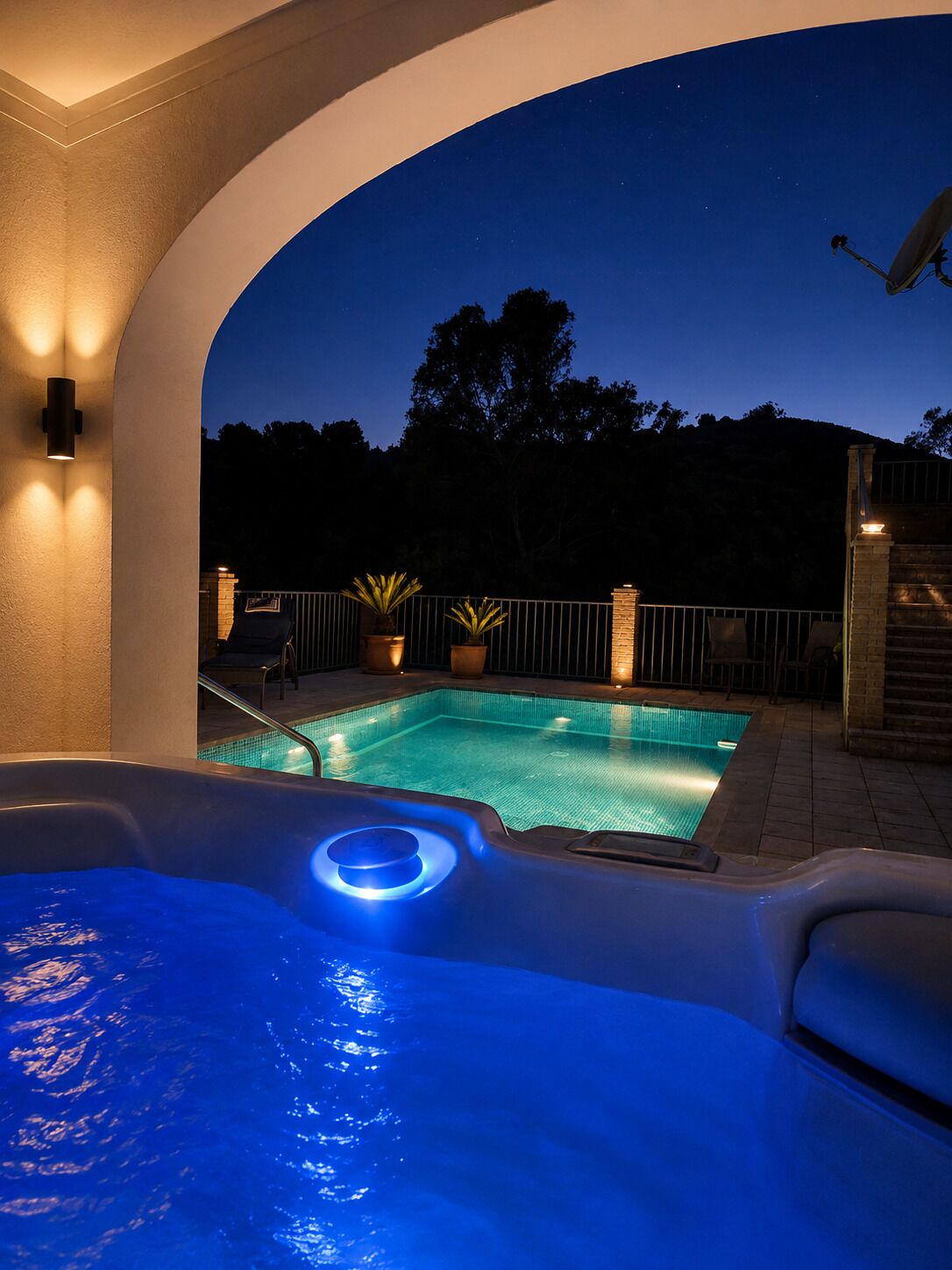 Ein Swimmingpool mit Whirlpool im Vordergrund, umgeben von einer Steinmauer und einem Metallzaun.