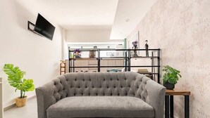 Un saló amb un sofà gris, una televisió penjada a la paret i un moble d'estanteries amb objectes decoratius.