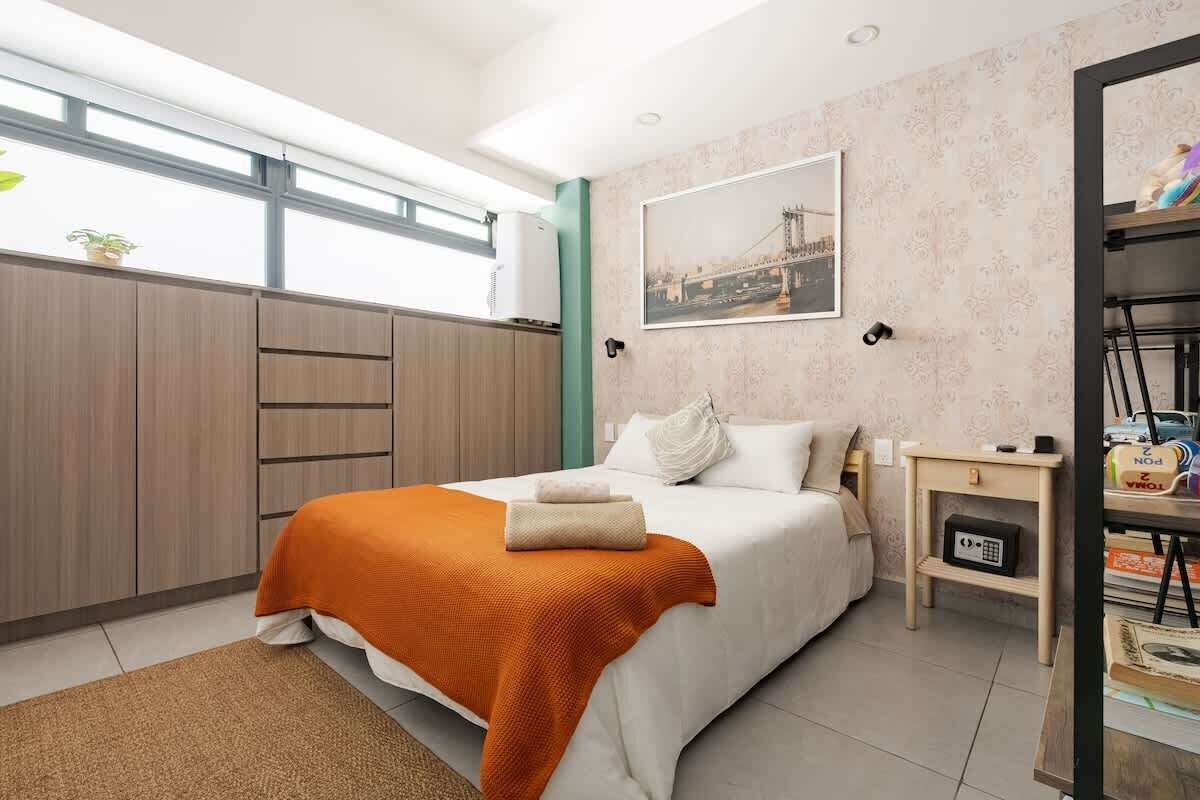 A hotel room with a bed, a nightstand, a wall-mounted air conditioner, a window with a view, and a framed picture on the wall.