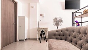 Habitació amb un sofà, un escriptori amb un tamboret, un armari, un ventilador i una televisió penjada a la paret.