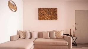 Uma sala de estar com um sofá seccional bege, um relógio de parede e uma obra de arte com textura.