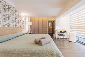 Quarto de hotel com cama, escrivaninha e cadeira. Há uma janela com persianas, uma luminária e uma porta que dá para outro quarto.