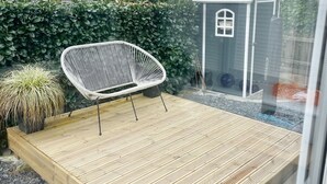 Une terrasse en bois avec une chaise blanche, un petit abri de jardin et une haie, le tout situé dans un quartier résidentiel.