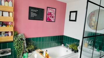 A bathroom with a white bathtub, green tiles, a pink wall, and a shelf with toiletries.