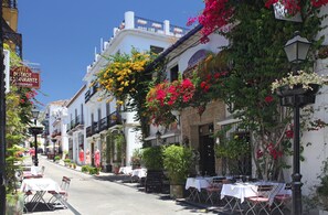 A street lined with white buildings, some with balconies and hanging flowers. There are tables and chairs outside, suggesting outdoor seating. - Casa Alma Marbella: private terrace, Wi-Fi & air conditioning (Marbella)