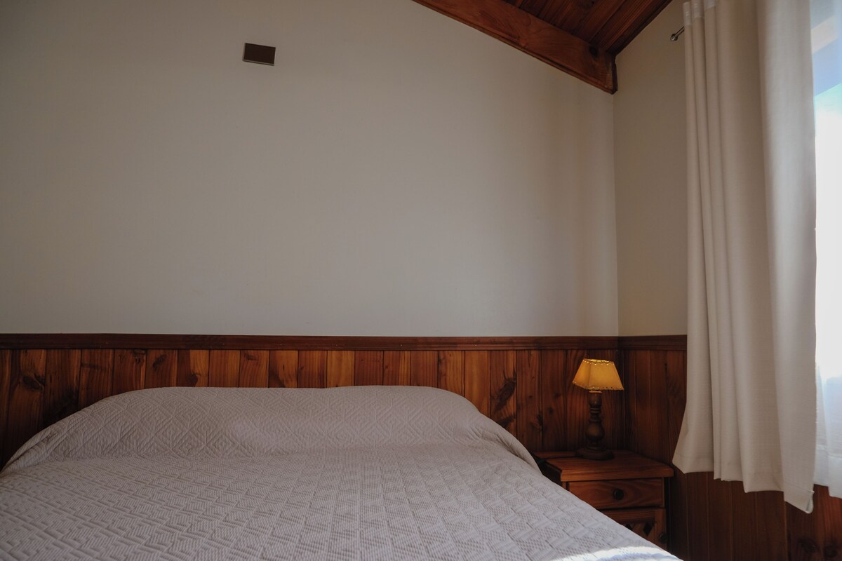 A bedroom with a wooden headboard, a single bed with a patterned bedspread, a nightstand with a lamp, and a window with curtains.