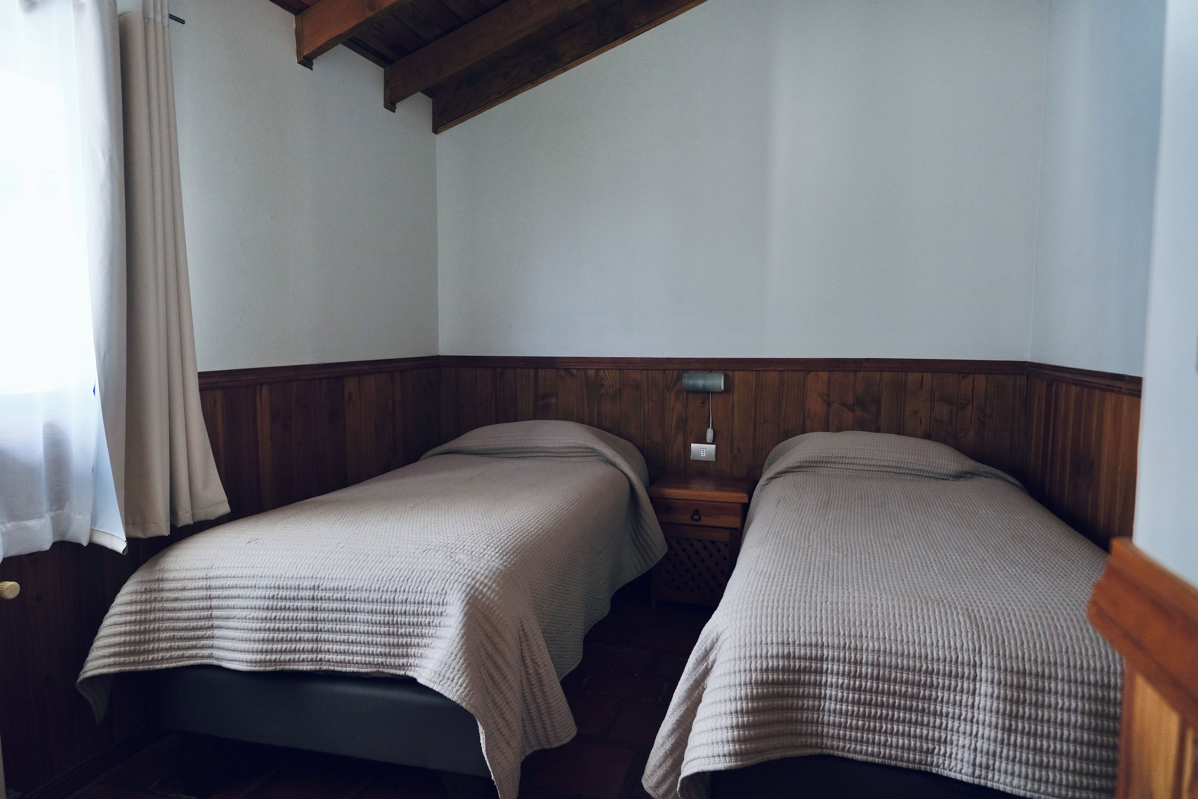 Una habitación con dos camas individuales, cabeceros de madera, una mesita de noche con lámpara y una ventana con cortinas.