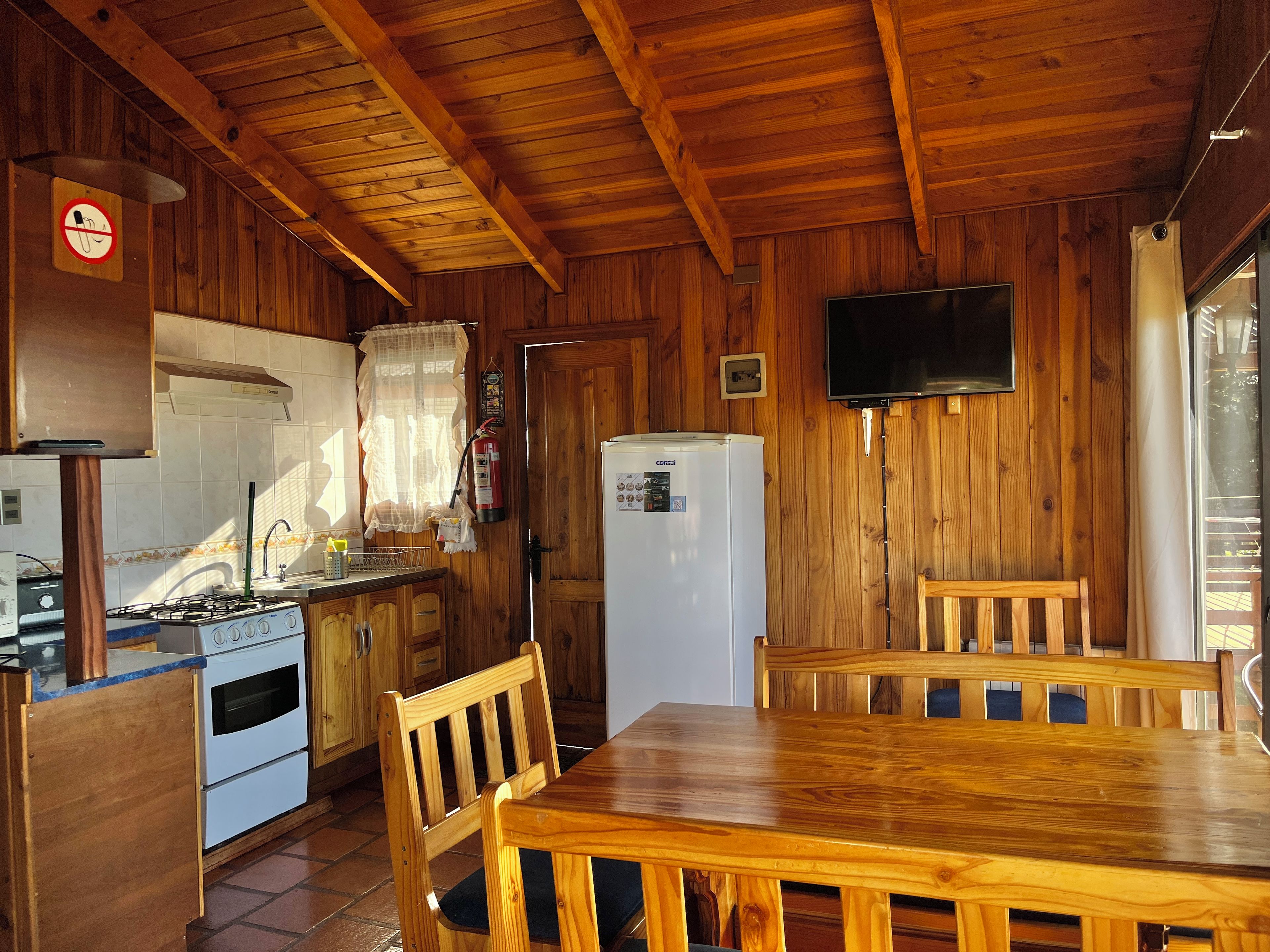 Un interior de madera con cocina, una mesa de comedor, sillas, un refrigerador y un televisor montado en la pared.