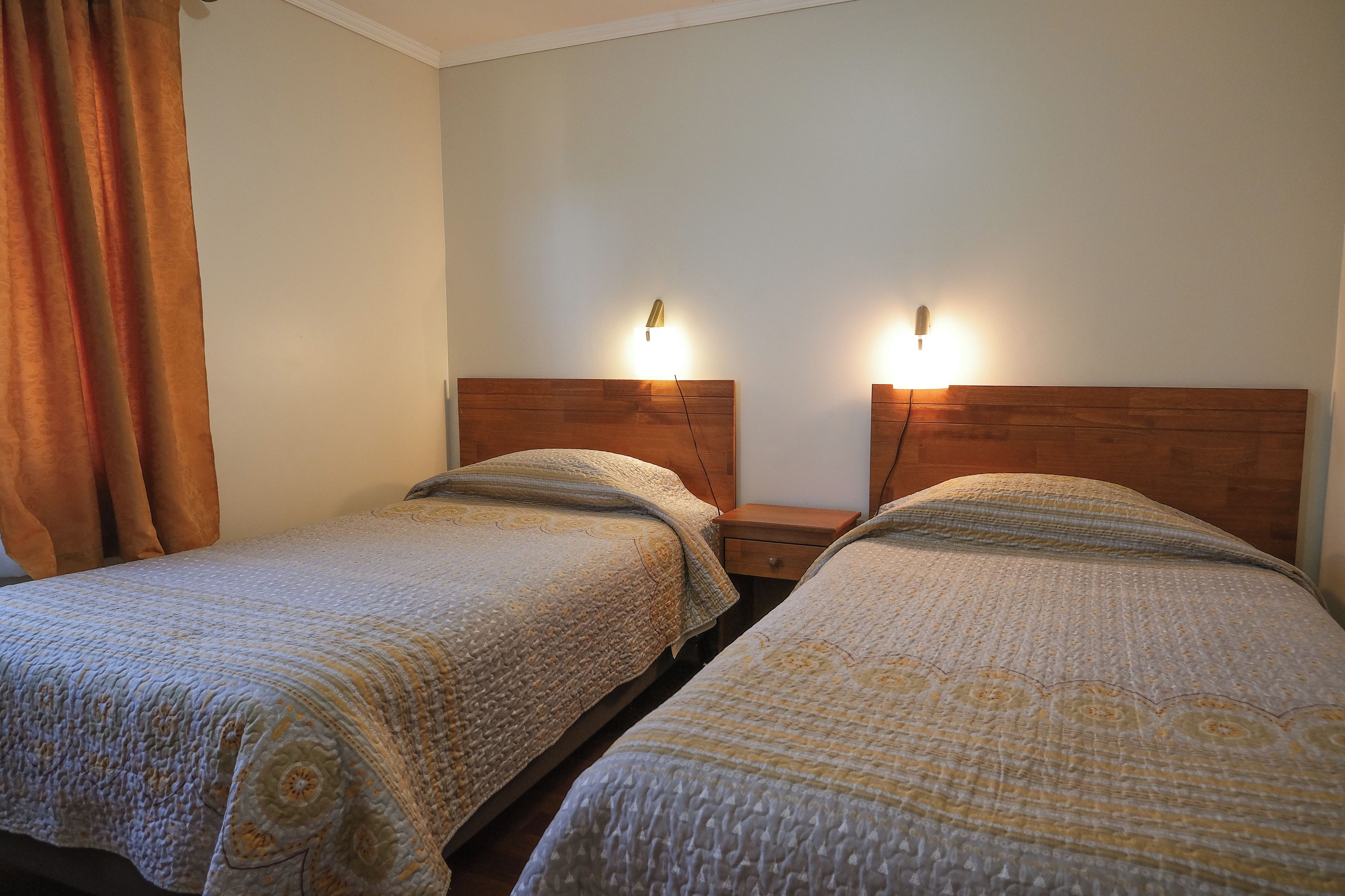 A hotel room with two single beds, a wooden headboard, a nightstand with a lamp, and a curtain.