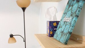 A room with a wooden shelf holding a tissue box and a decorative item, a floor lamp, and a towel on a table.