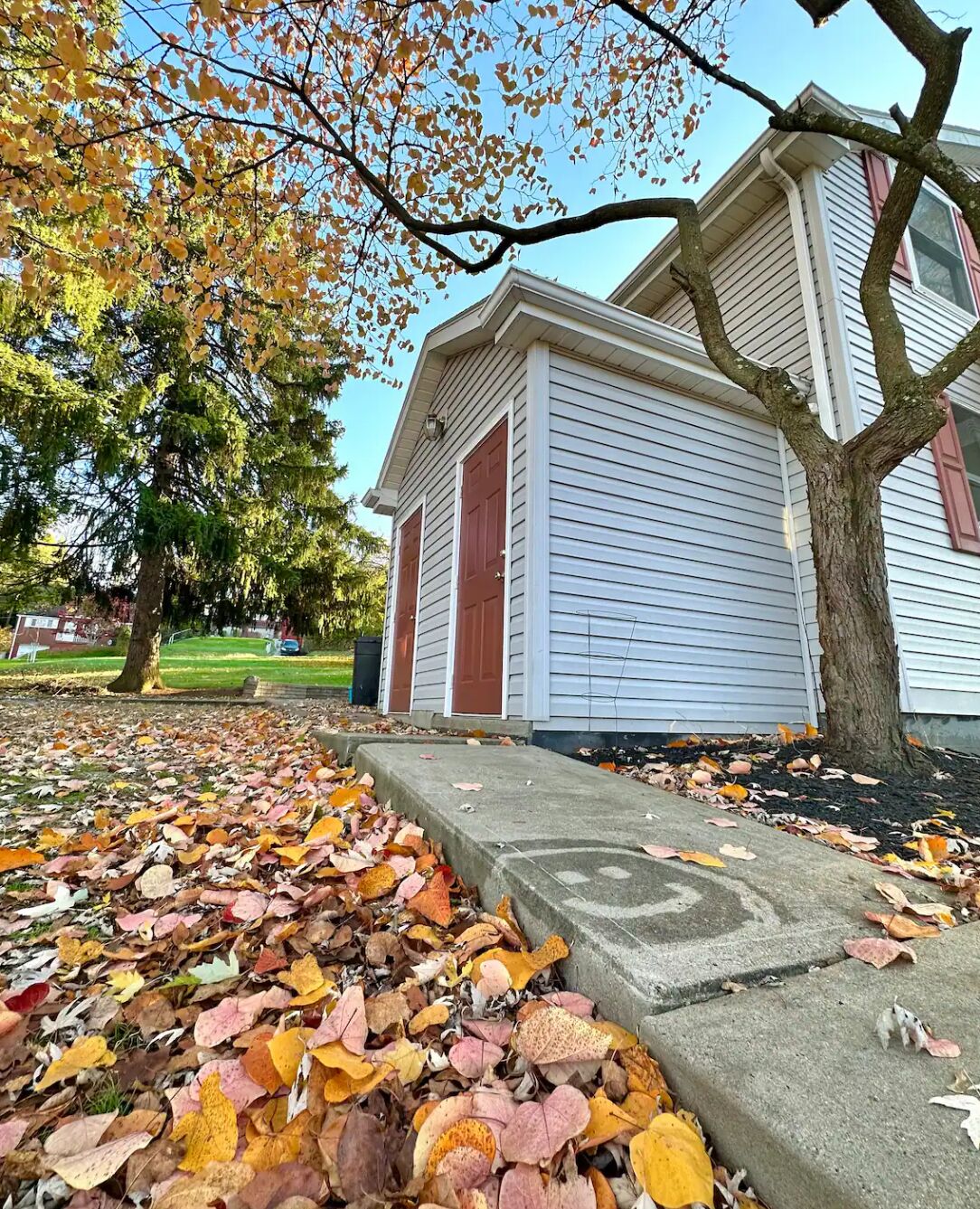 Een woonhuis met een veranda, een boom met herfstbladeren en een stoep vol gevallen bladeren.