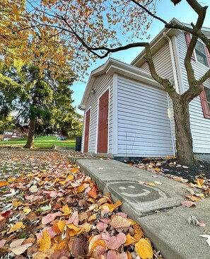 A residential building with a front porch, a tree with autumn leaves, and a sidewalk with fallen leaves.