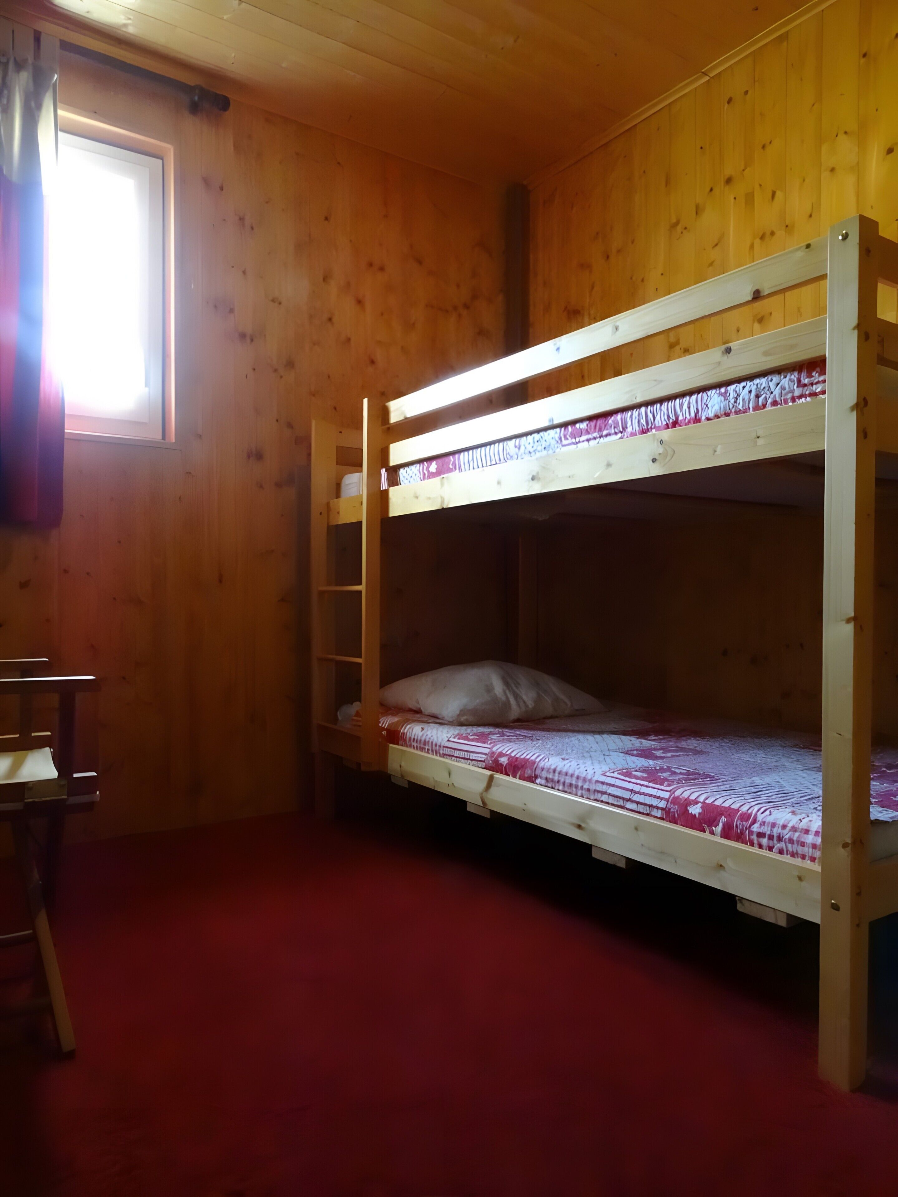 Ein Stockbett mit rotem Teppichboden, Holzwänden und einem Fenster mit Vorhängen.