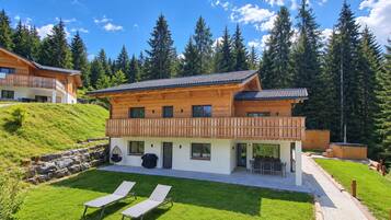 Ein zweistöckiges Holzhaus mit einem Balkon, einer Terrasse mit zwei Liegestühlen und einer Steinsichtmauer.