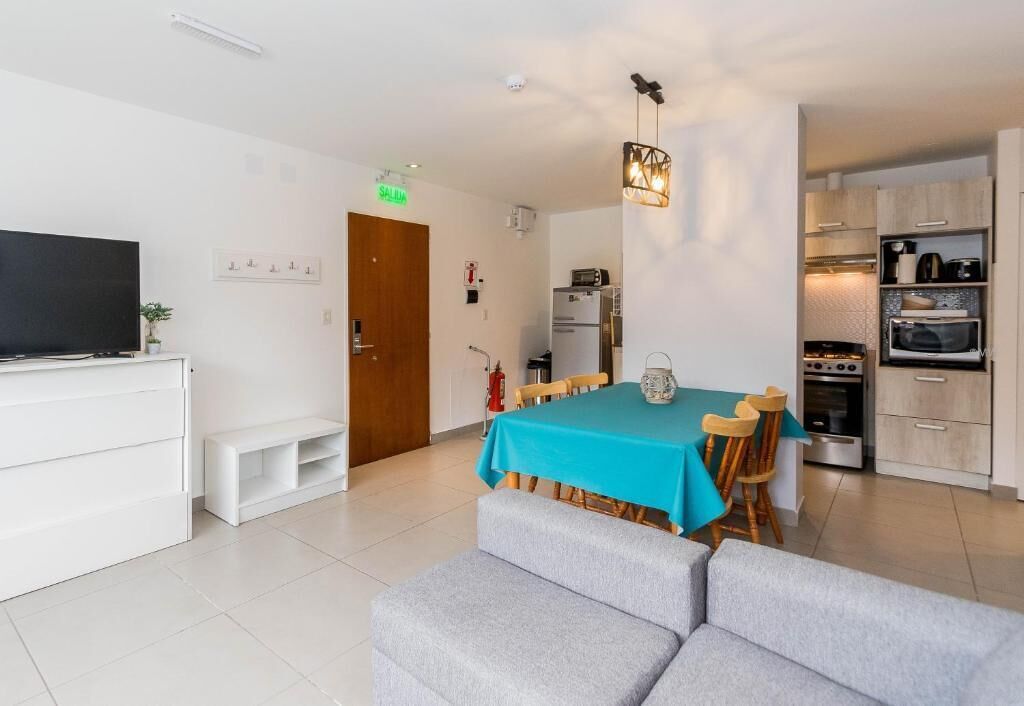 A modern living room with a flat-screen TV, a sofa, a dining table with chairs, and a kitchen area with appliances.