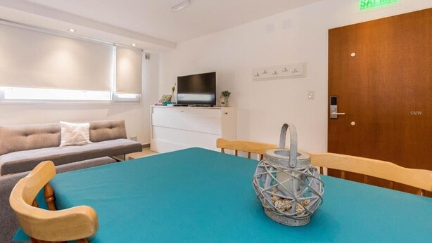 A hotel room with a sofa, a television on a dresser, a table with a blue tablecloth, and a door with a 'SALIDA' sign. - Australis Apartamento Micro-centro (Ushuaia)